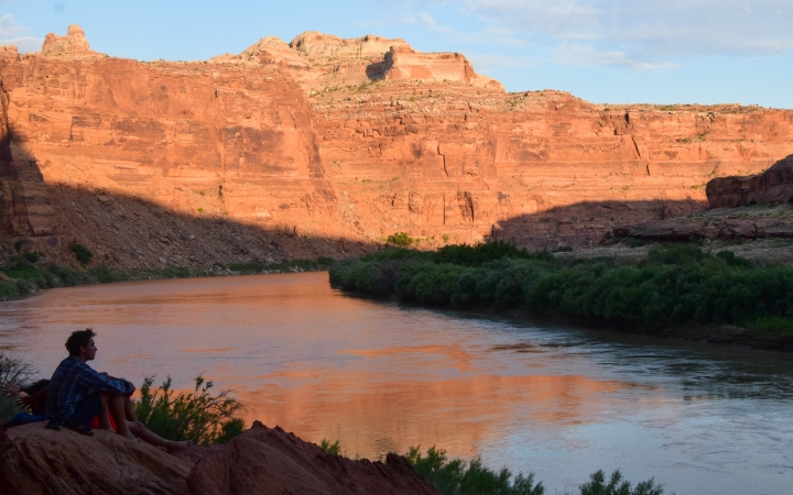 A person sits on the bank of a river, looking out over the water. In the background, there are tall red canyon walls.
